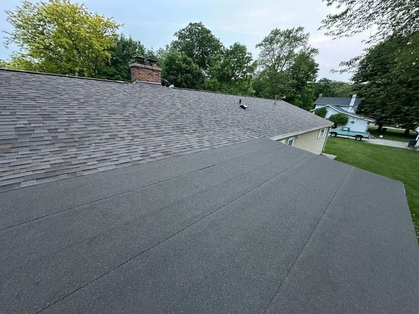 Residential roof with gray shingles and chimney, viewed from above, surrounded by trees.